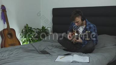 他在练习吉他。 吉他手在现代家庭的床上学习电吉他的新和弦。 耳机中的音乐家